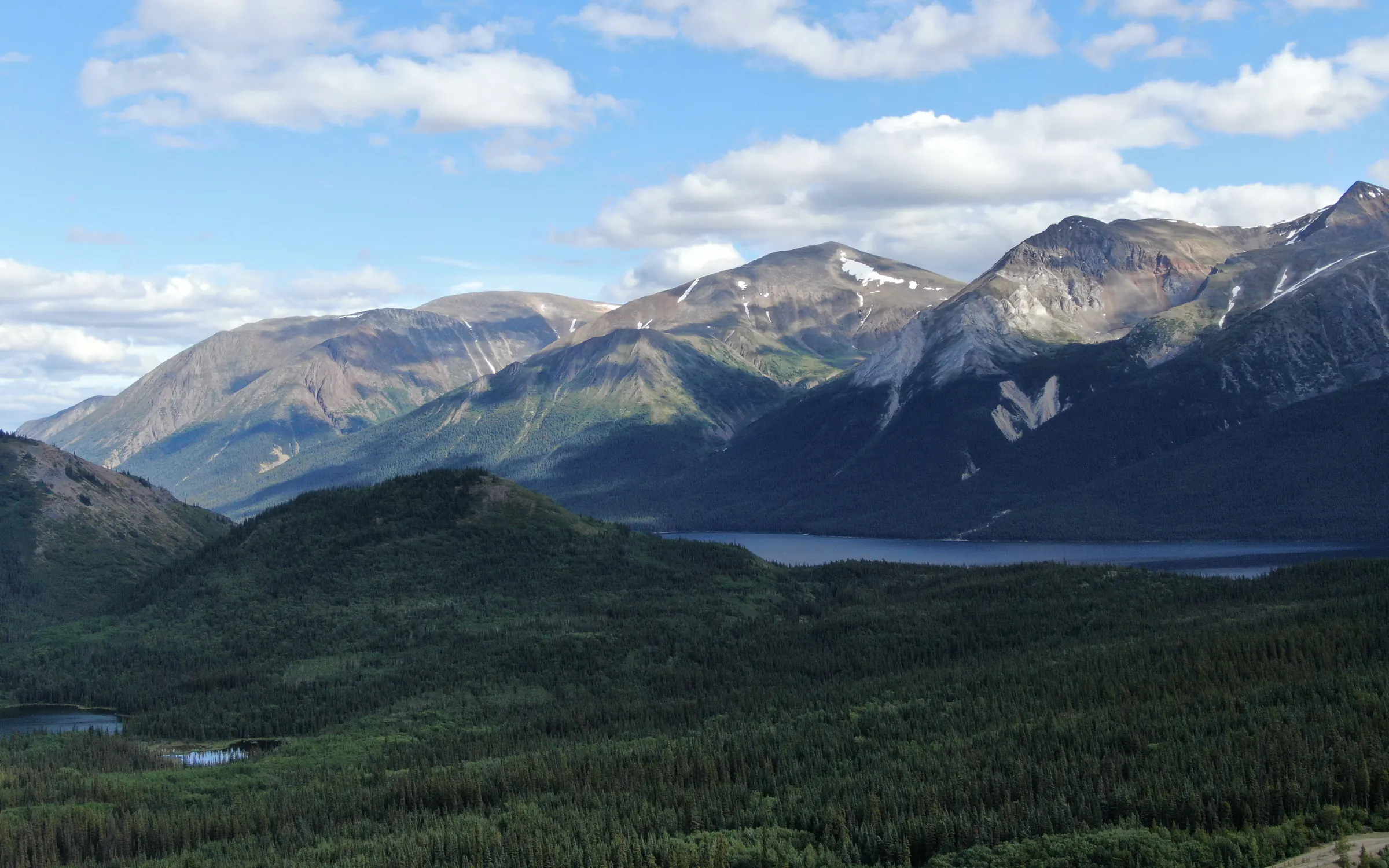 A mountain range above a lake and spruce forest, the kind of ground a drill program has to be aimed across
