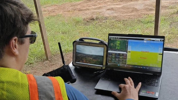 A Rosor pilot at a field ground station, watching survey telemetry on a laptop and the aircraft's camera feed on a rugged tablet