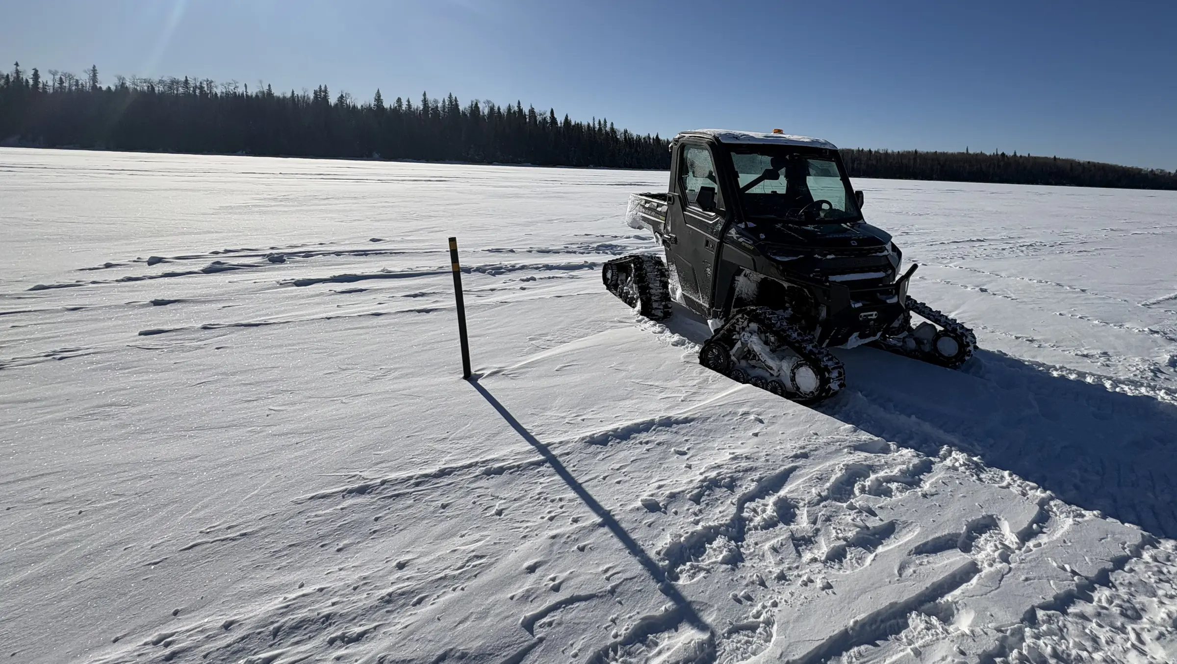 A tracked utility vehicle on a frozen lake at a Rosor survey site, with survey marker and crew tracks in the snow