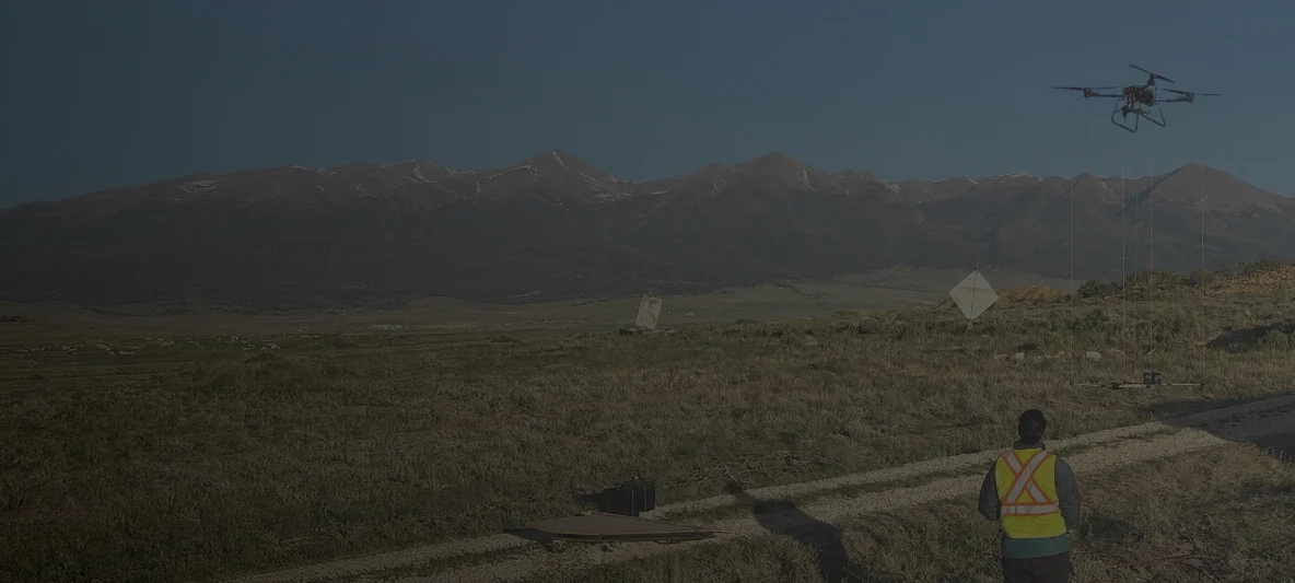 A Rosor survey drone hovering with its sensor slung beneath it on long lines, and the pilot in hi-vis on the ground below watching it, on open ground with a mountain range behind